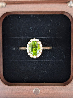 An oval green peridot ring with a halo of small white stones and a gold band, displayed on a black velvet insert inside a wooden box.