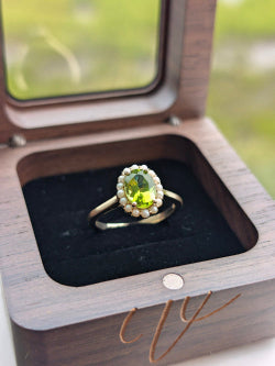A gold ring with a green peridot center stone and a halo of small clear stones, displayed in an open wooden jewelry box with a black velvet lining.